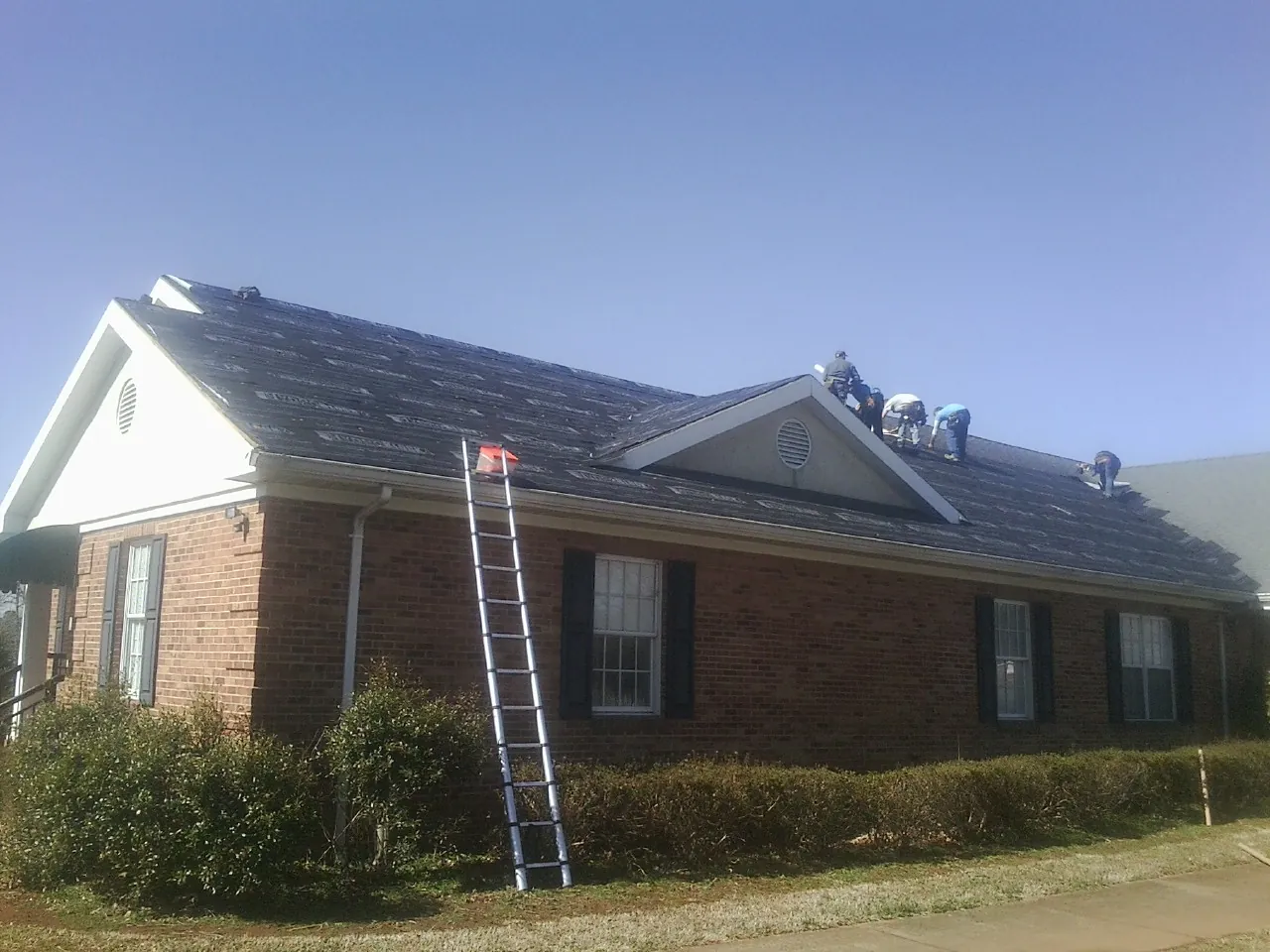 Crew tearing shingles off a brick ranch roof with a ladder set — Emergency Roof Repair work by our crew in Lovejoy